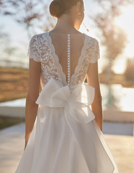Robes de mariées  ROSA CLARA AMEL : Robe de mariée de coupe évasée et style simple, confectionnée en tissu gazar et réhaussée de dentelle et pierreries. Elle offre un décolleté plongeant, un dos boutonné, des manches courtes et un nœud d’ornement dans le dos. Une délicate tenue 