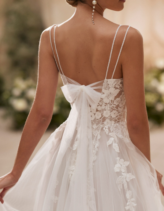 Robes de mariées  ADRIANA ALIER HERAIZ Robe de mariée longue et romantique en tulle, avec des détails en dentelle et perles. Elle présente un décolleté en V, un nœud décoratif dans le dos et une jupe fendue à l’avant. 