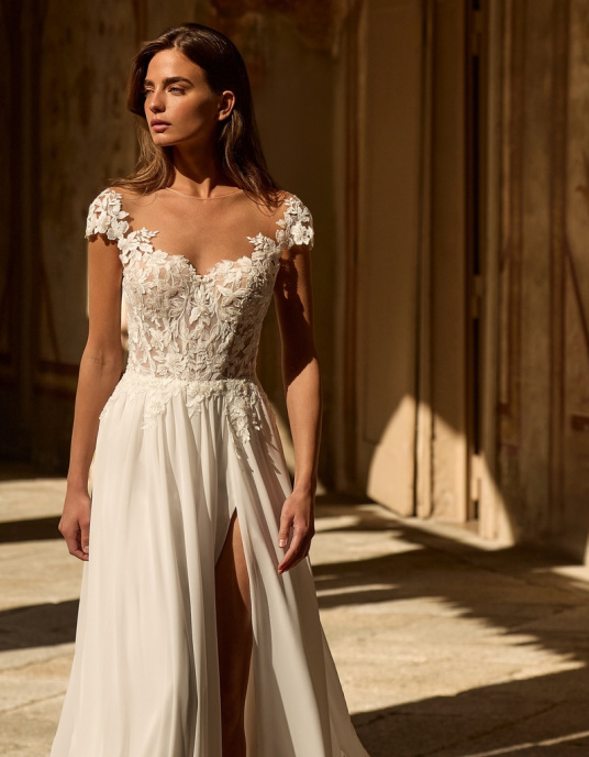 Robes de mariées  ALBERTO PALATCHI ADELE Robe de mariée trapèze de style romantique. Elle présente un corsage structuré en dentelle et perles, avec un décolleté illusion, sur une jupe en mousseline fendue sur le côté et ornée de délicates applications de dentelle. 