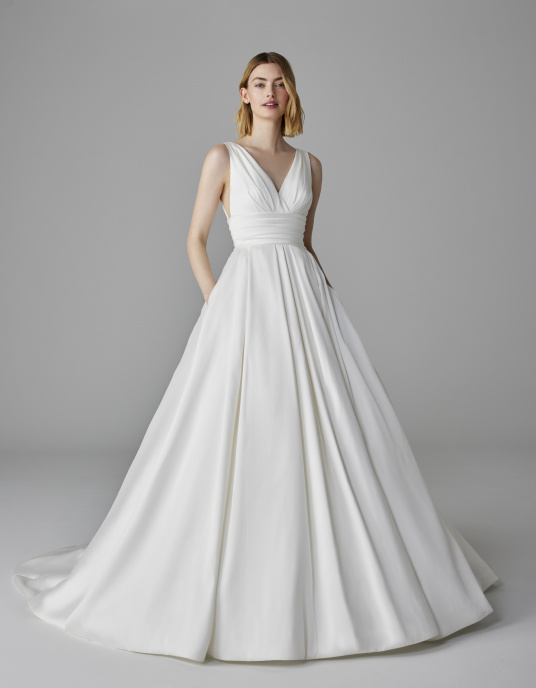 Robes de mariées  Pronovias LUMINEUSE-Cette robe en mikado présente des fentes en V sur le décolleté, sur les côtés et dans le dos, une ceinture large plissée et une superbe jupe princesse texturée ornée de poches pratiques. 