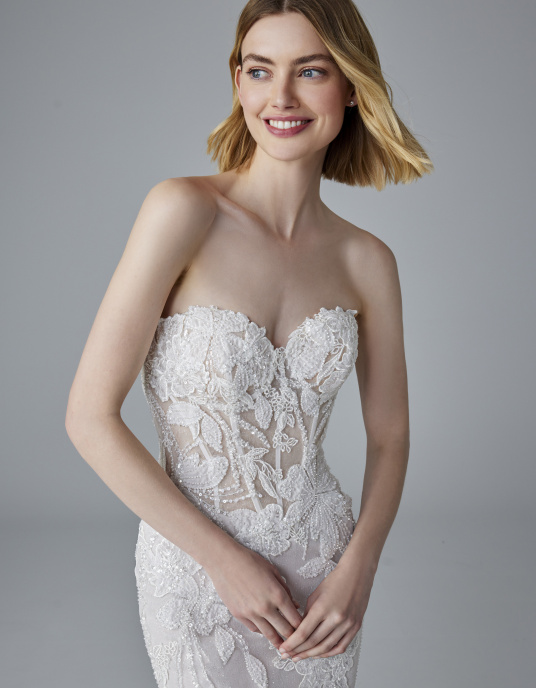 Robes de mariées  Pronovias NUAGE-un corsage renforcé style corset et une jupe seyante qui épouse la silhouette jusqu’aux genoux puis s’évase pour former une traîne sophistiquée. La dentelle et les strass créent des motifs et des formes qui font de ce modèle une véritable œuvre d’ar 