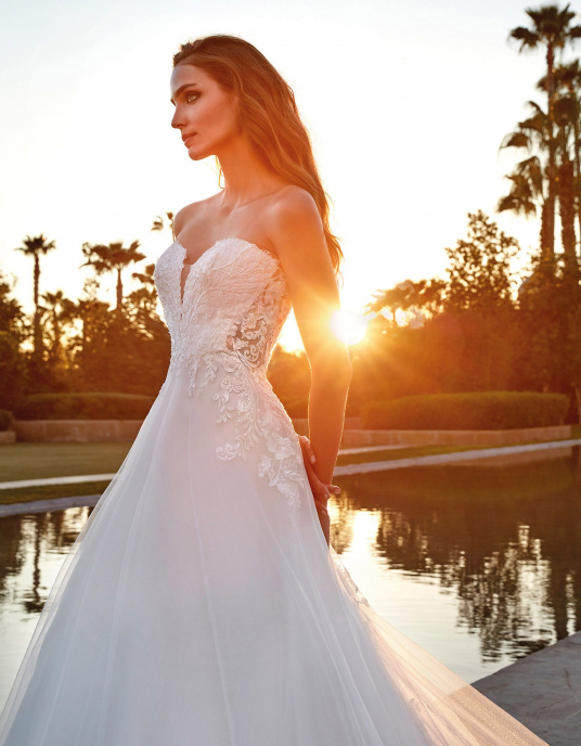 Robes de mariées  Pronovias RINNES Robe de mariée somptueuse au style princier. Le corsage sans manches, avec un décolleté en V, épouse parfaitement le corps, se prolongeant harmonieusement dans une jupe en A, ornée d'une surjupe en dentelle transparente et d'une traîne festonnée gl 